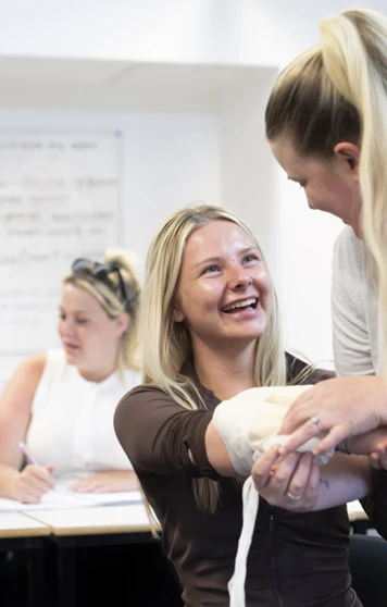 A student sits at a classroom desk, holding open a first aid manual with a bandage and tablet nearby, ready to tackle some hands-on learning A student sits at a classroom desk, holding open a first aid manual with a bandage and tablet nearby, ready to tackle some hands-on learning